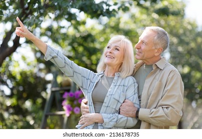 Happy Retired Couple Enjoying Time Together In Their Garden, Walking At Warm Spring Day, Man Embracing His Wife And Woman Pointing Aside, Showing Something. Let The Love Last Forever