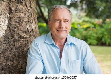 Happy Retired Man Sitting On Tree Trunk In The Park Looking At Camera