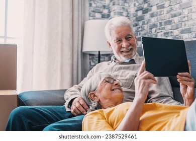 Happy retired senior family couple relaxing on sofa after moving activities, sharing tablet computer, looking at screen, laughing, making video call, using online app, Internet service - Powered by Shutterstock