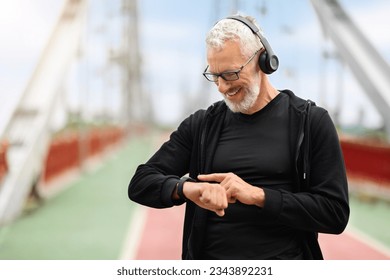 Happy retired sportsman exercising on footbridge, checking smart watch, copy space. Stopwatch, positive elderly man and runner check time, heart rate or monitor healthy body progress on data app - Powered by Shutterstock