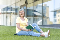 Happy Retired Woman Sitting On The Grass Resting On A Summer Day And Reading A Book, Looking At The Camera And Smiling