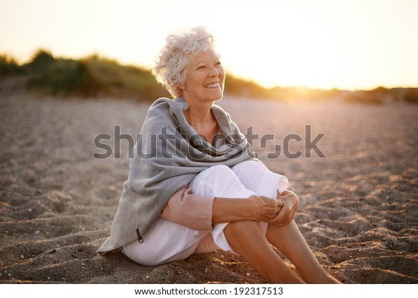 Happy retired woman wearing shawl sitting\
relaxed on sand at the beach. Senior caucasian woman sitting on the\
beach outdoors