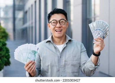 Happy Rich Asian Middle Aged Man Holding Two Fans Of Money, Celebrating Big Financial Luck And Success, Posing Outdoors. Profit And Wealth, Huge Lottery Win Concept