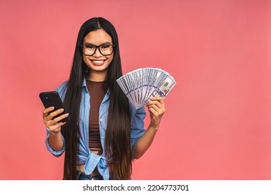 Happy Rich Winner! Portrait Of Asian Woman Holding Money Dollars Bills And Smiling Satisfied, Standing Isolated Over Pink Background. Using Mobile Phone.