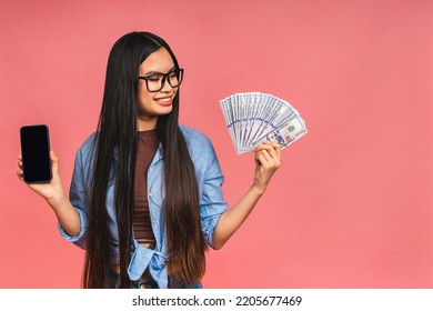 Happy Rich Winner! Portrait Of Asian Woman Holding Money Dollars Bills And Smiling Satisfied, Standing Isolated Over Pink Background. Using Mobile Phone. Showing Screen.