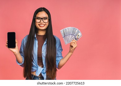 Happy Rich Winner! Portrait Of Asian Woman Holding Money Dollars Bills And Smiling Satisfied, Standing Isolated Over Pink Background. Using Mobile Phone. Showing Screen.