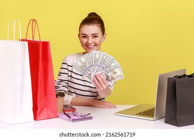 Happy Rich Woman Holding Out Money, Freelancer Showing Us Dollar Bills, Sitting At Workplace With Laptop And Shopping Bags. Indoor Studio Studio Shot Isolated On Yellow Background.