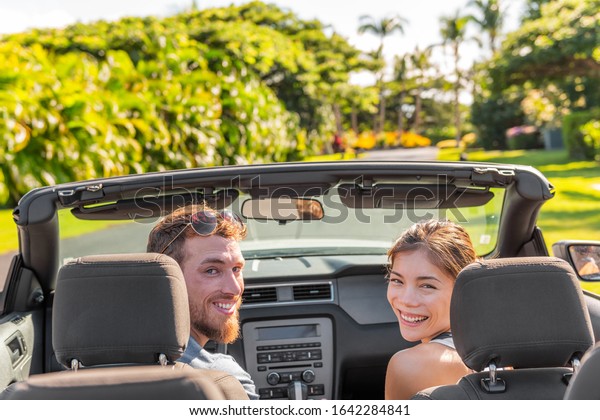 Happy road trip couple having fun driving luxury\
sports convertible car rental on travel vacation summer holidays\
looking back smiling. Interracial people tourists Asian woman,\
Caucasian man friends.