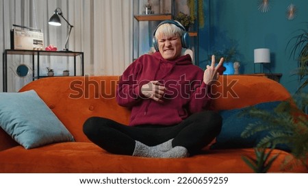 Happy rocker woman with short hair in wireless headphones relaxing at home dancing on couch listening energetic disco dancing music, playing on imaginary guitar. People weekend leisure activities