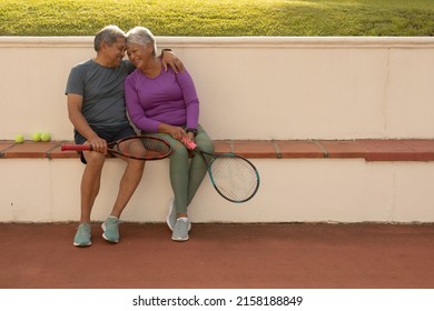 Happy romantic biracial senior couple holding tennis rackets while sitting on bench at tennis court. copy space, unaltered, sport, togetherness, love, retirement, healthy and active lifestyle. - Powered by Shutterstock