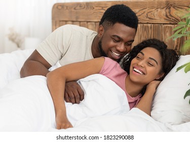 Happy Romantic Black Couple Laughing And Cuddling In Bed At Home, Empty Space. Emotional Young African American Lovers Playing In Bed, Man Hugging And Talking Sweet To His Joyful Girlfriend