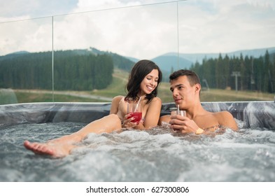 Happy romantic couple enjoying a bath in Jacuzzi while drinking cocktail outdoors on romantic vacation against blurred background of nature at the summer resort