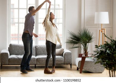 Happy Romantic Couple Having Fun In New Home, Man Holding Womans Hand Up Leading In Dance Enjoying Weekend, Cheerful Young Family Feeling Excited Spending Time Together In Cozy Modern Living Room