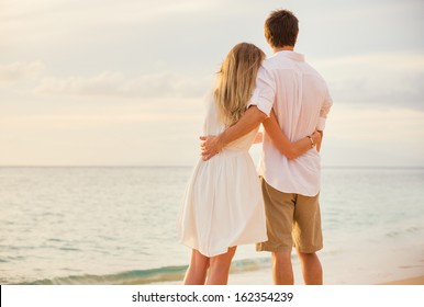 Happy romantic couple on the beach at sunset embracing each other. Man and woman in love watching the sun set into ocean
