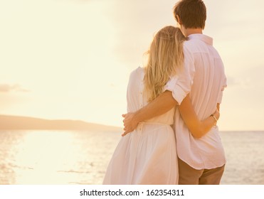 Happy romantic couple on the beach at sunset embracing each other. Man and woman in love watching the sun set into ocean