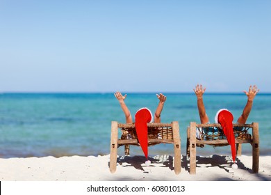 Happy Romantic Couple In Red Santa Hats At Tropical Beach Relaxing On Sun Beds