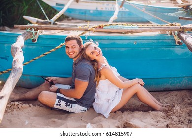 Happy Romantic Honeymoon Couple On The Beach Sitting On The Sand Near Old Fishing Boat And Looking Each Other Kissing. Husband With His Wife Near The Ocean. Sri Lanka. Hikkaduwa.