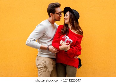 Happy Romantic Moment Of Two White People N Love Celebrating New Year Or Valentine’s Day.  Long Haired Woman With Gift Box In Hands Laughing. Wearing Trendy Red Knitted Sweater. Yellow Background.