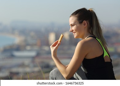 Happy Runner Woman Eating Energy Bar Sitting Outdoors After Exercise In City Outskirts