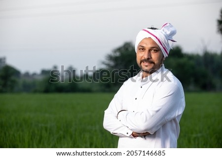 Happy rural Indian mustache man farmer giving side pose standing in field with hands crossed wearing kurta, smiling village male looking on camera in farm, greenery, blur background, copy space.