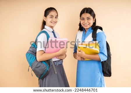 Happy Rural and Modern Indian student schoolgirls wearing school uniform holding books and bag standing together isolated over beige background, Studio shot, Education concept.