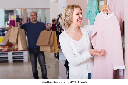 Happy Russian Elderly Wife Buying Dress At Apparel Store, Man Is Bored