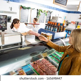 Happy Saleswoman Giving Meat Package To Female Customer At Counter In Butcher's Shop