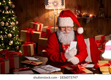 Happy Santa Claus Holding Smartphone Holding Cell Phone Using Mobile App For Social Distance Communication, Watching Videos Or Video Calling In Virtual Online Chat Sit At Home Table Late On Xmas Eve.