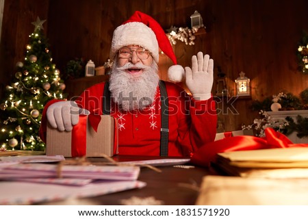 Happy Santa Claus, Saint Nicholas holding letter looking to camera, talking to webcam video calling or recording Happy New Year, Merry Christmas greetings on xmas eve sitting at table late at home.