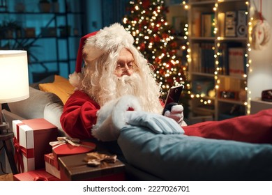 Happy Santa Claus sitting on the couch at home and using his smartphone, he is connecting online and sending virtual wishes - Powered by Shutterstock