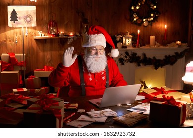 Happy Santa Claus waving hand video calling kid talking to child greeting on Merry Christmas, Happy New Year in virtual video online chat on laptop sitting at home table late with present on xmas eve.