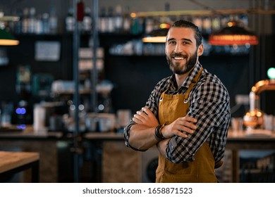 Happy satisfied bartender with apron near counter with crossed arms and looking at camera. Confident coffee shop waiter smiling at cafeteria. Successful proud young brewer at his pub with copy space. - Powered by Shutterstock