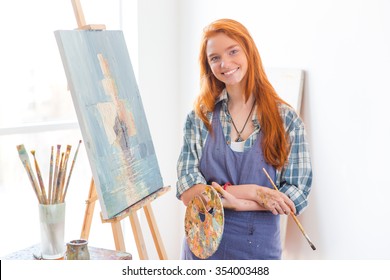 Happy Satisfied Beautiful Young Woman Painter In Apron Finished Painting Picture In Art Studio