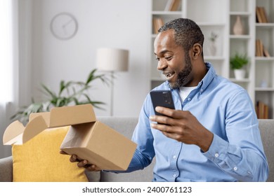 Happy and satisfied buyer received cardboard box in mail, satisfied with speed of delivery of a gift for an online store, an African American man with a phone in hands confirms receipt of the goods. - Powered by Shutterstock