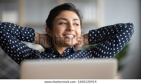 Happy satisfied indian woman rest at home office\
sit with laptop hold hands behind head, dreamy young lady relax\
finished work feel peace of mind look away dream think of future\
success concept