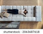 Happy satisfied lazy Asian woman relaxing on big comfortable couch, enjoying comfort, leisure on weekend, sleeping, napping, breathing fresh conditioned air in living room. Top view, full length