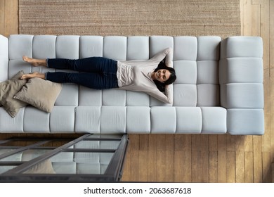 Happy satisfied lazy Asian woman relaxing on big comfortable couch, enjoying comfort, leisure on weekend, sleeping, napping, breathing fresh conditioned air in living room. Top view, full length