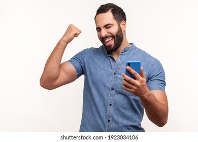 Happy satisfied man with beard in blue shirt holding smartphone and smiling making yes gesture, celebrating online lottery or giveaway victory. Indoor studio shot isolated on white background