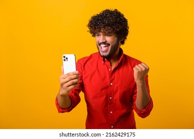 Happy satisfied man with beard holding smartphone and smiling making yes gesture, celebrating online lottery or giveaway victory. Indoor studio shot isolated on orange background