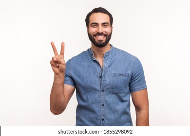 Happy Satisfied Man Showing V Sign Symbol Of Peace With Fingers, Looking At Camera With Toothy Smile, Celebrating His Successful Victory. Indoor Studio Shot Isolated On White Background