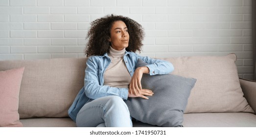 Happy satisfied relaxed young african woman lounge on comfortable modern furniture sofa. Smiling black girl sitting on couch in cozy living room looking away, dreaming, feel no stress at home concept.