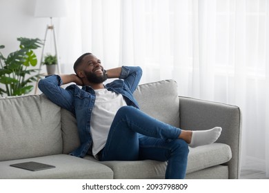 Happy Satisfied Relaxed Young Black Bearded Male Resting In Living Room Interior With Tablet On Sofa. Break After Work In Own Apartment, Thinking About New Business Ideas At Home, Self-isolation