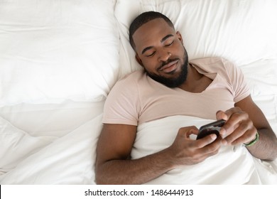 Happy satisfied rested african american man lying at comfortable bed on soft pillow in bedroom at home or hotel, waking up, using mobile phone, playing online game, reading sms from beloved woman.
