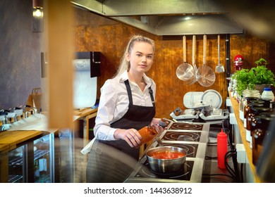 Happy Satisfied Woman Adding Condiments For Taste In Boiling Pot With Pasta Sauce – Small Business Woman Looking At Camera And Cooking In Restaurant Kitchen - Chef In Apron Preparing Food Recipe