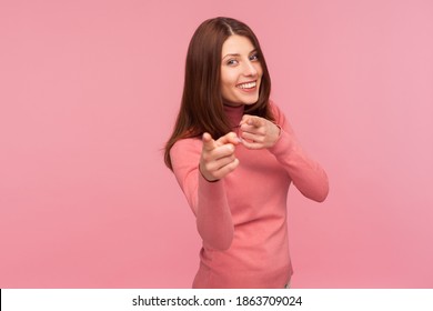 Happy satisfied woman with brown hair in pink sweater pointing fingers on you looking at camera with toothy smile, congratulating with victory. Indoor studio shot isolated on pink background