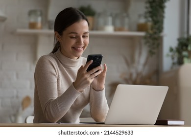 Happy satisfied young entrepreneur, business woman using mobile phone at home workplace, chatting, browsing internet, reading message with good news, sitting at table with laptop in kitchen