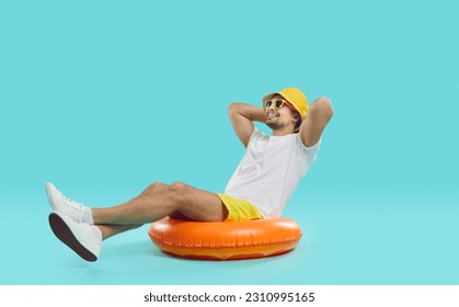 Happy satisfied young man enjoys summer sea vacation sitting on inflatable swimming circle. Joyful guy in summer outfit sits on inflatable circle with his hands behind his head on blue background