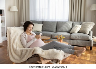 Happy Satisfied Young Pregnant Woman Shopping Online From Home, Using Smartphone For Buying Baby Clothes, Consulting Doctor On Virtual Chat. Expecting Mom With Cellphone Relaxing In Armchair