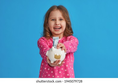 Happy savings. A little cute girl with a piggy bank on a blue background. The child smiles happily, puts the money in the piggy bank and holds it out. The concept of saving money for a dream. - Shutterstock ID 1945079728
