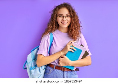 happy school girl with bag and books posing at camera isolated over purple background, cheerful female with curly hair smile, happy to go school. education concept